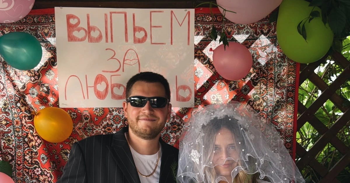Жених в тёмных очках и невеста в фате у ковра с плакатом «Выпьем за любовь»