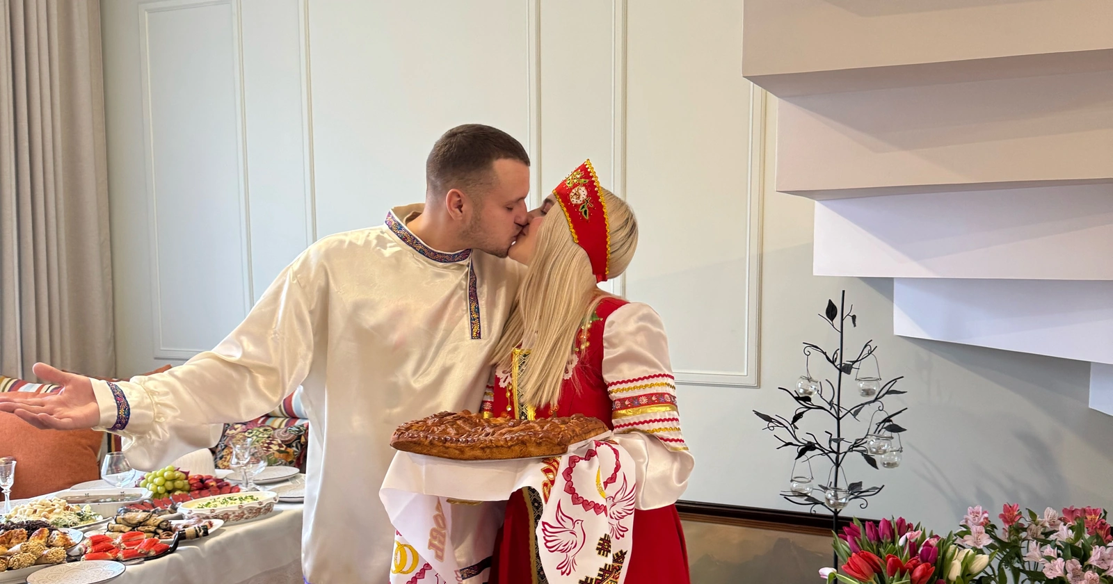 Пара в русских костюмах целуется дома, у невесты в руках пирог на рушнике