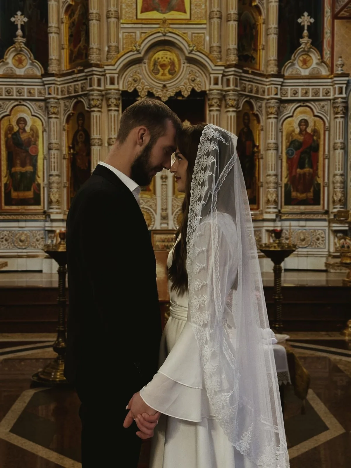 Жених и невеста стоят лбом ко лбу перед иконостасом, невеста в кружевном платке