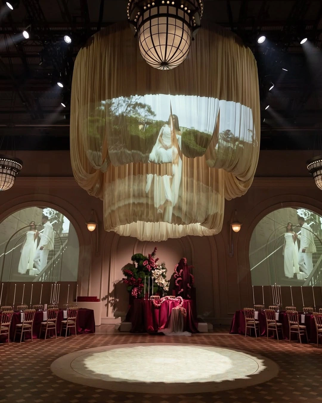 Банкетный зал с драпированной люстрой и бордовыми столами перед праздником