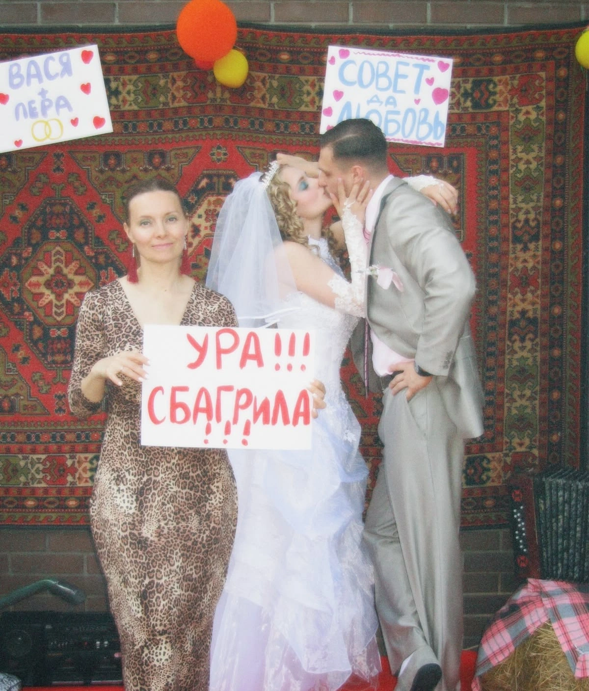 Подруга невесты с плакатом «Ура, сбагрила» у ковра, молодые целуются рядом